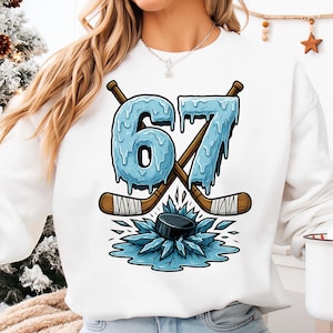 May include: White sweatshirt with a hockey graphic. The design shows the number "67" in a dripping ice effect, crossed hockey sticks, and a hockey puck on ice. The graphic is in shades of blue, brown, and black.
