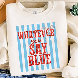 May include: Cream-colored sweatshirt with the text "WHATEVER you SAY BLUE" in red letters, set against a backdrop of vertical light blue stripes. A small red bow is at the top. The sweatshirt is a casual style.