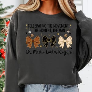May include: Dark gray sweatshirt with the text "CELEBRATING THE MOVEMENT, THE MOMENT, THE MAN" and "Dr. Martin Luther King Jr." Three decorative bows in brown, black, and cream colors are featured below the text.