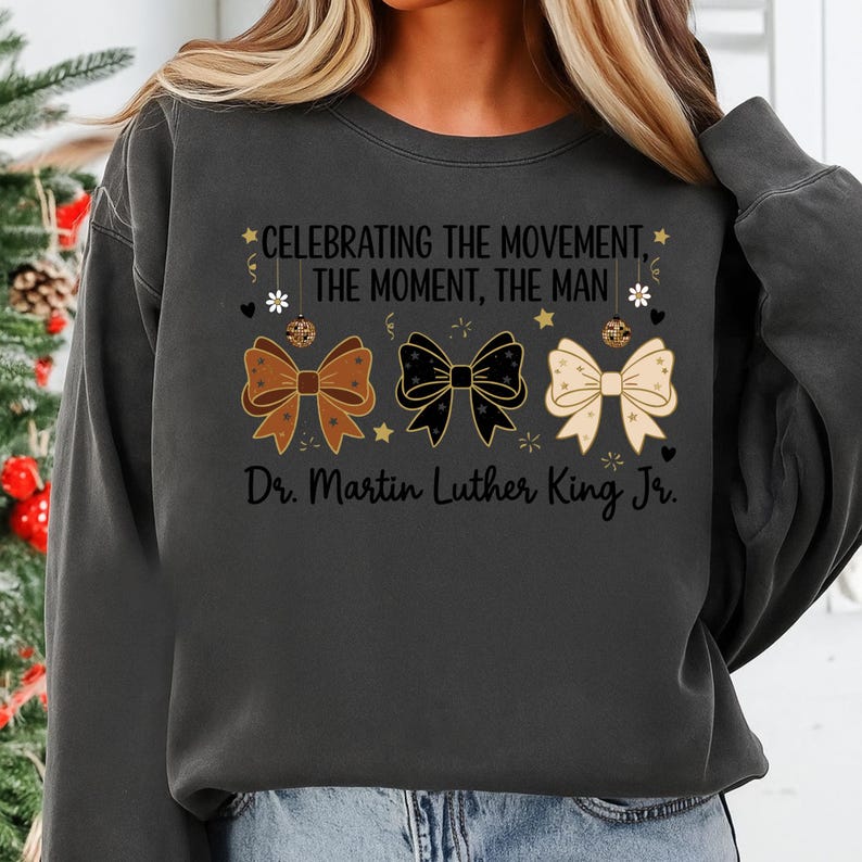 May include: Dark gray sweatshirt with the text "CELEBRATING THE MOVEMENT, THE MOMENT, THE MAN" and "Dr. Martin Luther King Jr." Three decorative bows in brown, black, and cream colors are featured below the text.