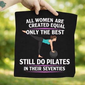 Op de afbeelding: Zwarte draagtas met de tekst "ALL WOMEN ARE CREATED EQUAL ONLY THE BEST STILL DO PILATES IN THEIR SEVENTIES". De afbeelding toont een vrouw die Pilates doet met een bal. De achtergrond is wazig groen.