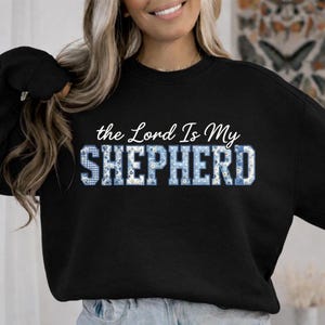 Puede incluir: Sudadera negra con la frase "the Lord Is My SHEPHERD". La palabra "SHEPHERD" presenta un diseño floral azul y blanco. Una prenda superior casual y cómoda.