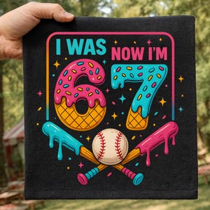 Puede incluir: Toalla negra con el texto "I WAS NOW I'M 67" en un diseño colorido. Los números están diseñados para parecerse a donas y helados. Una pelota de béisbol y bates cruzados en la parte inferior.