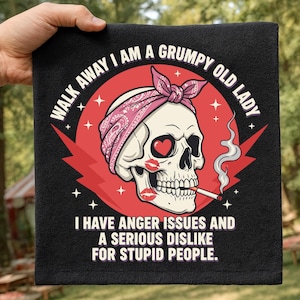 Puede incluir: Toalla negra con un diseño de calavera con un pañuelo rosa y marcas de lápiz labial. El texto dice "WALK AWAY I AM A GRUMPY OLD LADY" y "I HAVE ANGER ISSUES AND A SERIOUS DISLIKE FOR STUPID PEOPLE."