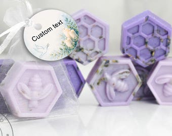 May include: Light purple hexagonal soaps featuring embossed bee and honeycomb patterns with embedded dried lavender bits. One soap is wrapped in a sheer white organza bag with a round white tag attached by a ribbon, displaying the text Custom text.