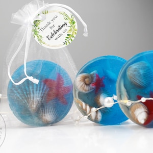 May include: Circular blue soaps with seashells and starfish encased within. One soap is in a white organza bag with a tag saying "Thank you for Celebrating with us." A pearl string and the words "Sandra Soap Handmade soap favors" are also visible.