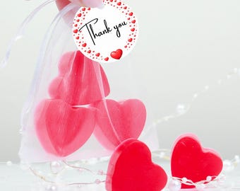 May include: Red heart-shaped soaps in a sheer bag, with a "Thank you" tag. Several soaps are visible, some inside the bag and others arranged nearby. A string of pearls adds a decorative touch. The soaps are a Valentine's Day or wedding favor.