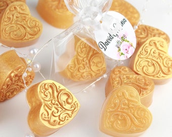 May include: Golden heart-shaped soaps with ornate designs, some individually wrapped in clear cellophane. A tag reads "David & Anna with love" with a floral accent. The soaps are arranged with pearl-like beads and a string of lights.