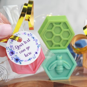 May include: Three hexagonal shaped soaps in a clear plastic bag with a gold ribbon. The soaps are green, light green, and orange. The green soap has a honeycomb pattern, the light green soap has a bee design, and the orange soap has a honeycomb pattern. A white circle tag with purple flowers and the text "Your text can go here" is attached to the bag.