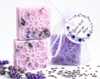 May include: Two square light purple soap bars with an intricate geometric cutout pattern and embedded dried lavender buds. One bar is inside a sheer white organza drawstring bag with a round tag featuring a floral wreath and the text Your text can go here.