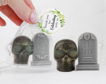 May include: A sheer white organza drawstring bag containing a gray tombstone soap with the text IGNORE THE SCREAMS and a metallic bronze skull soap. Beside it sits a matching metallic bronze skull soap and a gray tombstone soap labeled BE BACK.
