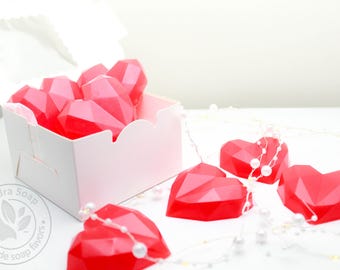 May include: A white box with a scalloped edge holds several faceted, heart-shaped soaps in shades of red. Several more soaps are scattered around the box, some adorned with strings of pearls. The soaps are handmade, as indicated by the logo.