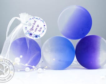 May include: Three round bars of purple and white ombre soap arranged with a white organza drawstring bag. A circular tag attached to the bag features a floral border and the text Your text can go here. A string of small white faux pearls is present.