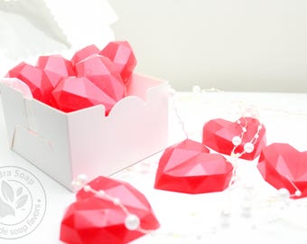 May include: A white cardboard box filled with multiple bright red geometric heart-shaped soaps. Additional red heart-shaped soaps are arranged outside the box, intertwined with a delicate string of small white pearls. A circular logo reads Sandra Soap Handmade soap favors.