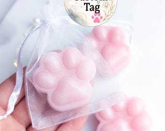 May include: Three light pink paw-shaped soaps. One soap is inside a small white organza drawstring bag. A circular tag above features the text Custom Tag with pink paw prints and floral line art. The soaps have a smooth, glossy finish.