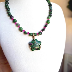 Könnte beinhalten: Eine Edelsteinkette, die auf einer weißen Schaufensterpuppe ausgestellt ist. Die Kette hat grüne und rosa Perlen und einen sternförmigen Anhänger. Der Anhänger ist ein grüner Stein mit rosa Einschlüssen, umwickelt mit goldenem Draht.