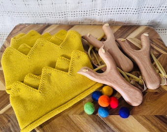 Puede incluir: Tres coronas de punto color amarillo mostaza con borde superior en zigzag, tres tirachinas de madera con bandas elásticas y seis pequeñas bolas de fieltro en colores verde, azul, naranja, rojo y azul oscuro sobre una superficie de madera.