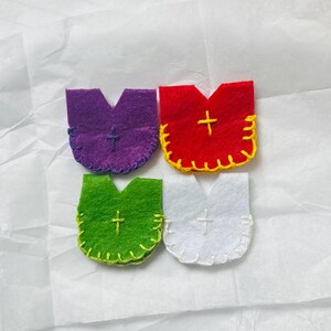 Puede incluir: Cuatro adornos de fieltro en morado, rojo, verde y blanco, cada uno con una cruz y costuras decorativas. Los adornos tienen una forma &uacute;nica, que se asemeja a una vestidura o a una letra estilizada.