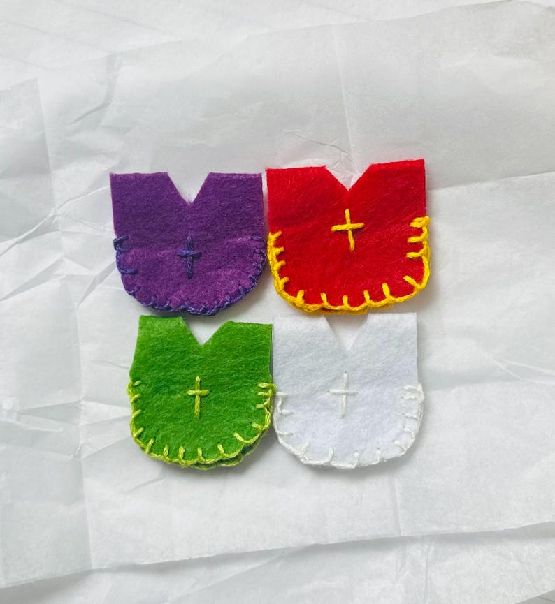 Puede incluir: Cuatro adornos de fieltro en morado, rojo, verde y blanco, cada uno con una cruz y costuras decorativas. Los adornos tienen una forma &uacute;nica, que se asemeja a una vestidura o a una letra estilizada.