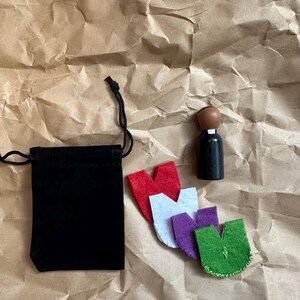 Puede incluir: Una bolsa de terciopelo negro con cord&oacute;n, un peque&ntilde;o tap&oacute;n de botella de madera y cuatro formas de fieltro en rojo, blanco, morado y verde est&aacute;n dispuestas sobre papel marr&oacute;n. El tap&oacute;n tiene una parte superior marr&oacute;n y una base negra.