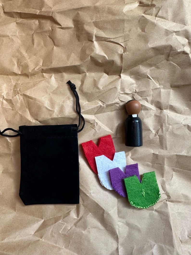 Puede incluir: Una bolsa de terciopelo negro con cord&oacute;n, un peque&ntilde;o tap&oacute;n de botella de madera y cuatro formas de fieltro en rojo, blanco, morado y verde est&aacute;n dispuestas sobre papel marr&oacute;n. El tap&oacute;n tiene una parte superior marr&oacute;n y una base negra.