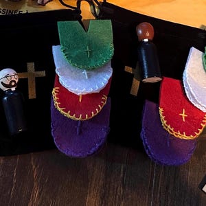 Puede incluir: Un surtido de art&iacute;culos de tem&aacute;tica religiosa, que incluyen vestiduras de fieltro en verde, blanco, rojo y morado, con una cruz dorada. Una mu&ntilde;eca de madera con gafas y un atuendo negro est&aacute; junto a una bolsa negra con una cruz dorada.