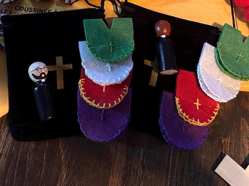 Puede incluir: Un surtido de art&iacute;culos de tem&aacute;tica religiosa, que incluyen vestiduras de fieltro en verde, blanco, rojo y morado, con una cruz dorada. Una mu&ntilde;eca de madera con gafas y un atuendo negro est&aacute; junto a una bolsa negra con una cruz dorada.