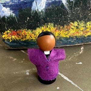Puede incluir: Mu&ntilde;eca de madera artesanal que representa una figura religiosa. La mu&ntilde;eca tiene una cabeza marr&oacute;n con cabello negro, un cuello blanco y una vestimenta de fieltro morado. El fondo presenta una pintura colorida de un paisaje.