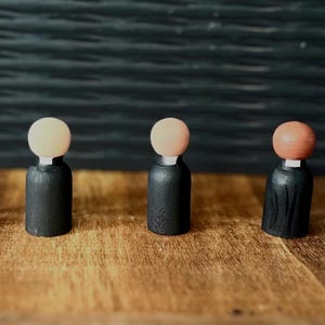 Puede incluir: Cinco mu&ntilde;ecos de madera con cuerpos negros y cabezas de color claro. Los mu&ntilde;ecos est&aacute;n dispuestos sobre una superficie de madera, con un fondo oscuro y texturizado. Las cabezas son de color beige claro o naranja.