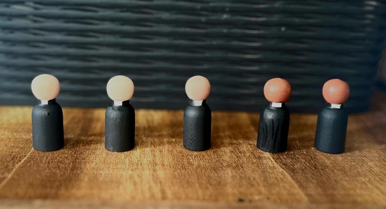 Puede incluir: Cinco mu&ntilde;ecos de madera con cuerpos negros y cabezas de color claro. Los mu&ntilde;ecos est&aacute;n dispuestos sobre una superficie de madera, con un fondo oscuro y texturizado. Las cabezas son de color beige claro o naranja.