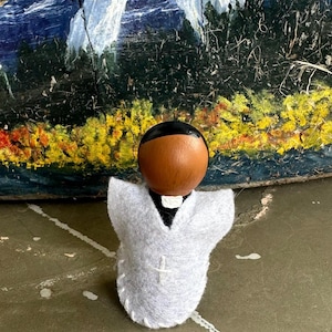 Puede incluir: Mu&ntilde;eca de madera artesanal que representa a un sacerdote, con cabeza marr&oacute;n y pelo negro. La mu&ntilde;eca lleva una t&uacute;nica de fieltro blanco con una cruz y un cuello negro. El fondo presenta una pintura abstracta colorida.