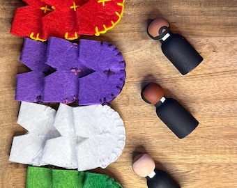 Puede incluir: Una bandeja de madera contiene formas de fieltro de colores y pequeñas figuras de madera. Las formas de fieltro son rojas, moradas, blancas y verdes, con costuras de cruz y costuras amarillas o verdes. Las figuras de madera son negras con cabezas marrones o tostadas.