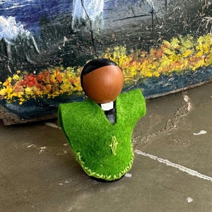 Puede incluir: Una mu&ntilde;eca de madera hecha a mano con cabeza marr&oacute;n y pelo negro, que lleva un chaleco de fieltro verde brillante. El chaleco est&aacute; ribeteado con costuras verdes y presenta un peque&ntilde;o dise&ntilde;o bordado. La mu&ntilde;eca est&aacute; sobre un fondo de madera pintada.