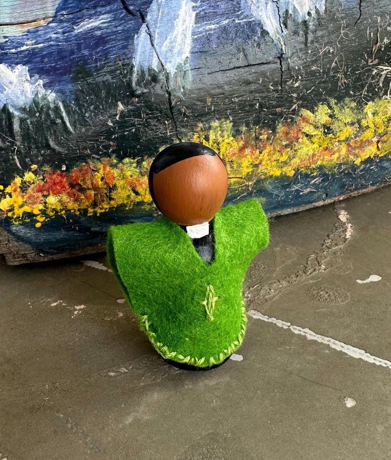 Puede incluir: Una mu&ntilde;eca de madera hecha a mano con cabeza marr&oacute;n y pelo negro, que lleva un chaleco de fieltro verde brillante. El chaleco est&aacute; ribeteado con costuras verdes y presenta un peque&ntilde;o dise&ntilde;o bordado. La mu&ntilde;eca est&aacute; sobre un fondo de madera pintada.