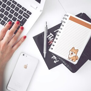 May include: A white notebook with a brown header and a cute cartoon Shiba Inu sticker. The notebook has a spiral binding and is lying on a black surface.