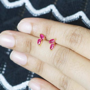 Puede incluir: Un par de pendientes de oro con tachuelas, cada uno con tres gemas rojas vibrantes dispuestas en un diseño de hoja. Los pendientes se muestran en una mano sobre un fondo con estampado en blanco y negro.