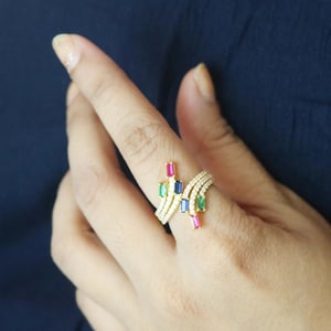 Könnte beinhalten: Ein goldfarbener Ring mit einem einzigartigen Design, besetzt mit rechteckigen Edelsteinen in Rot, Grün, Blau und Gelb. Der Ring ist außerdem mit kleinen, klaren Edelsteinen besetzt. Der Ring wird an einem Finger vor einem dunkelblauen Hintergrund getragen.