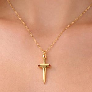 May include: A gold-coloured pendant necklace featuring a dagger design with red accents. The dagger pendant hangs from a delicate chain. The necklace is worn against a white background.