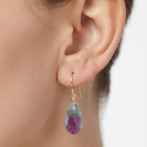 May include: A teardrop-shaped gemstone earring featuring a faceted surface with mottled shades of deep pink, green, and grey. The stone is suspended from a thin, curved gold-toned wire hook that passes through the earlobe for secure placement.