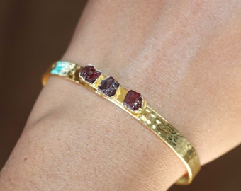 May include: Gold bangle bracelet with three red garnet stones set in the metal. The bracelet has a hammered texture.