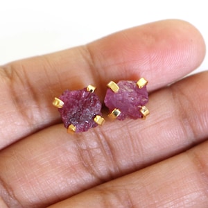 Puede incluir: Un par de pendientes de oro con piedras preciosas de rubí rosa. Las piedras preciosas son en bruto y tienen un aspecto áspero y natural.