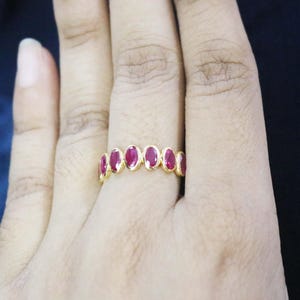 May include: A gold ring set with a row of oval-cut, deep red gemstones. The gemstones are closely set, forming a continuous band. The ring is shown on a hand, highlighting its design and craftsmanship. The ring is likely made of gold.
