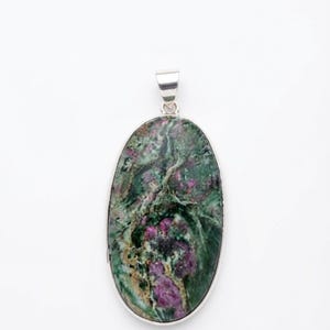 Puede incluir: Un colgante de forma ovalada con una piedra preciosa de rubí zoisita pulida engastada en un marco de metal plateado. La piedra muestra un patrón moteado de verde bosque profundo, negro e inclusiones de color magenta vibrante.
