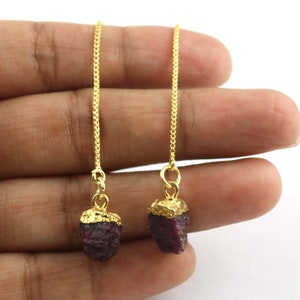 Puede incluir: Un par de pendientes de cadena dorada con piedras preciosas de rubí rojo oscuro en bruto. Los rubíes están cubiertos con metal dorado. Los pendientes se muestran sobre un fondo blanco.
