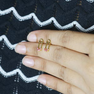 Puede incluir: Pendientes dorados con forma de imperdible y piedras rosas. Los pendientes son pequeños y delicados, con un diseño clásico de imperdible y una fila de piedras rosas en la parte inferior.