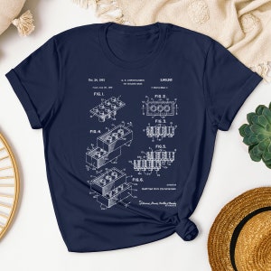 May include: A navy blue t-shirt with a white print of a patent drawing for a toy building brick. The patent drawing shows different views of the brick and includes the text "Oct. 24, 1961", "G. K. CHRISTIANSEN", "3,005,282", "FIG. 1", "FIG. 2", "FIG. 3", "FIG. 4", "FIG. 5", "FIG. 6", and "Godtfred Kirk Christiansen".