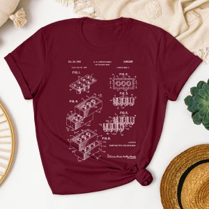 May include: A maroon t-shirt with a white print of the Lego patent, showing different configurations of the iconic building brick. The patent number is 3,005,282 and the date is Oct. 24, 1961.