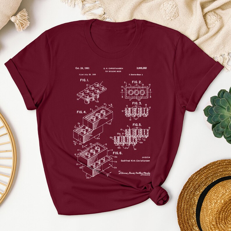 May include: A maroon t-shirt with a white print of the Lego patent, showing different configurations of the iconic building brick. The patent number is 3,005,282 and the date is Oct. 24, 1961.