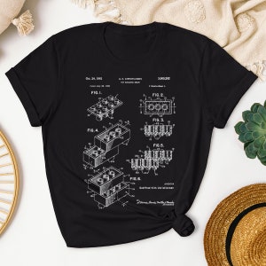 May include: A black t-shirt with a white print of the Lego patent, showing different views of the iconic building brick. The patent number is 3,005,282 and the date is Oct. 24, 1961. The text "Godtfred Kirk Christiansen" is also included.