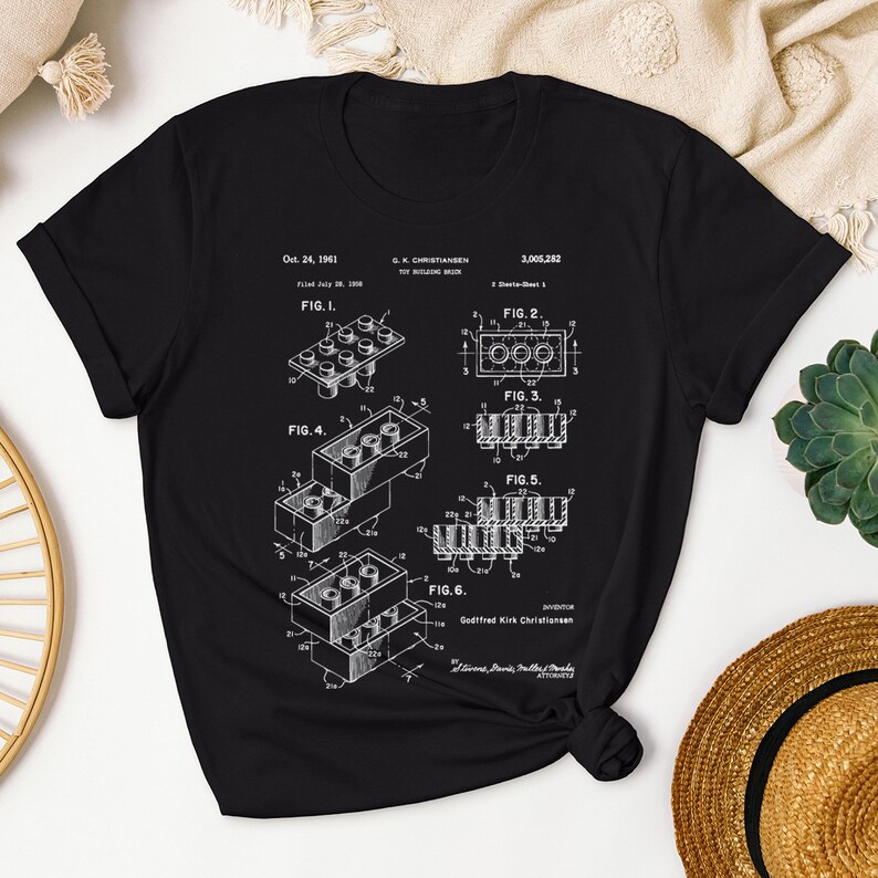 May include: A black t-shirt with a white print of the Lego patent, showing different views of the iconic building brick. The patent number is 3,005,282 and the date is Oct. 24, 1961. The text "Godtfred Kirk Christiansen" is also included.